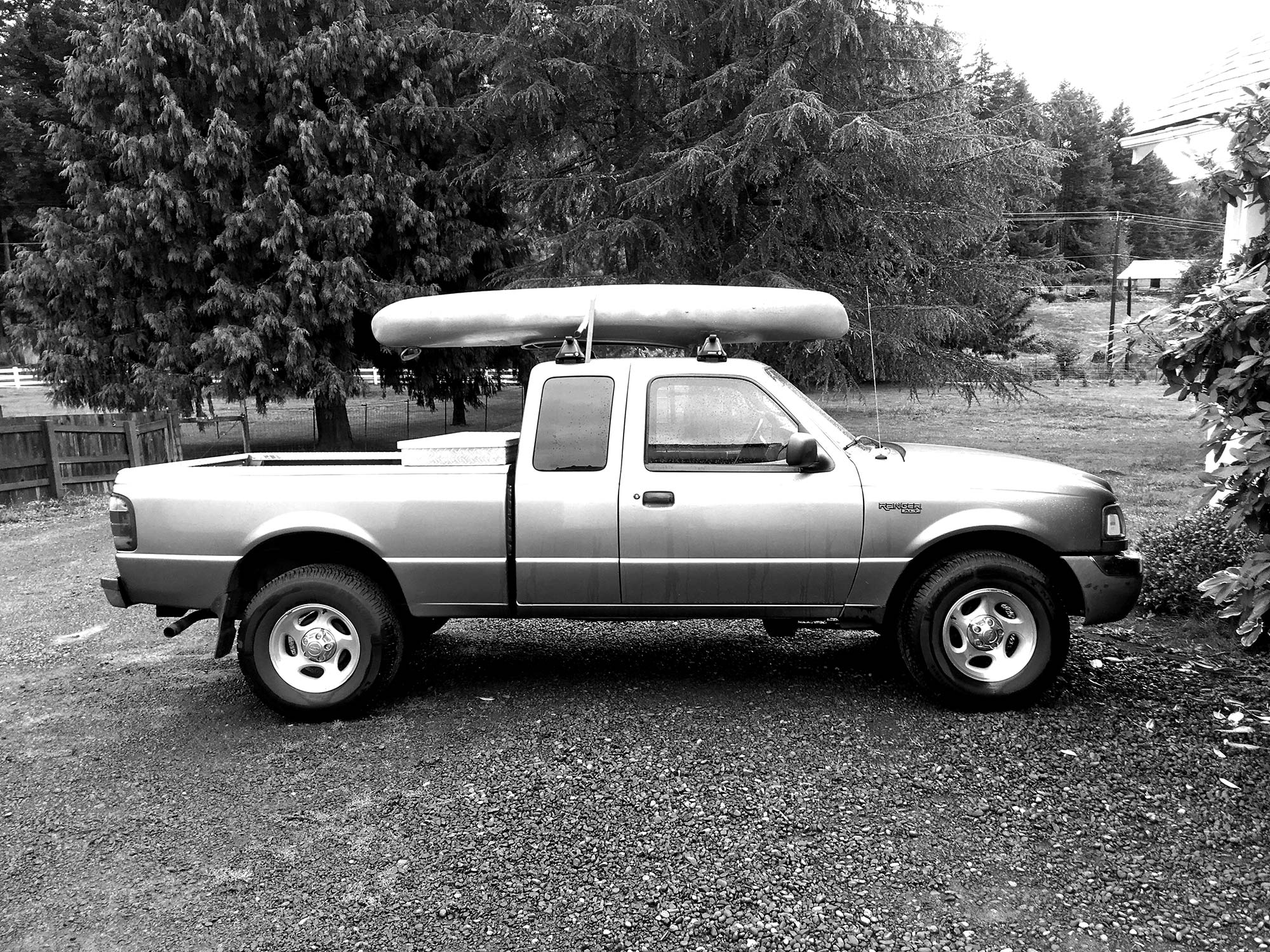 Black and white photo of the ford ranger with a kayak on top in a gravel driveway with a big tree in the background