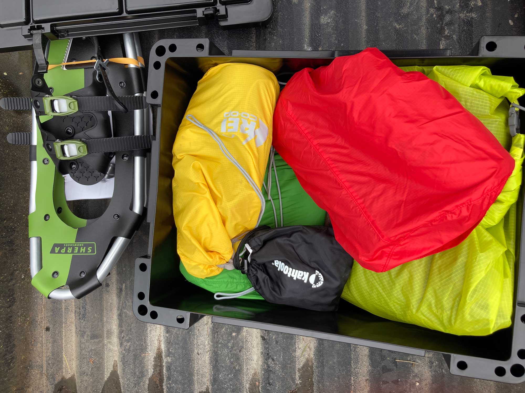 Snowshoes at left in a truck bed, with a black box full of kit bags in green, red, yellow, and black