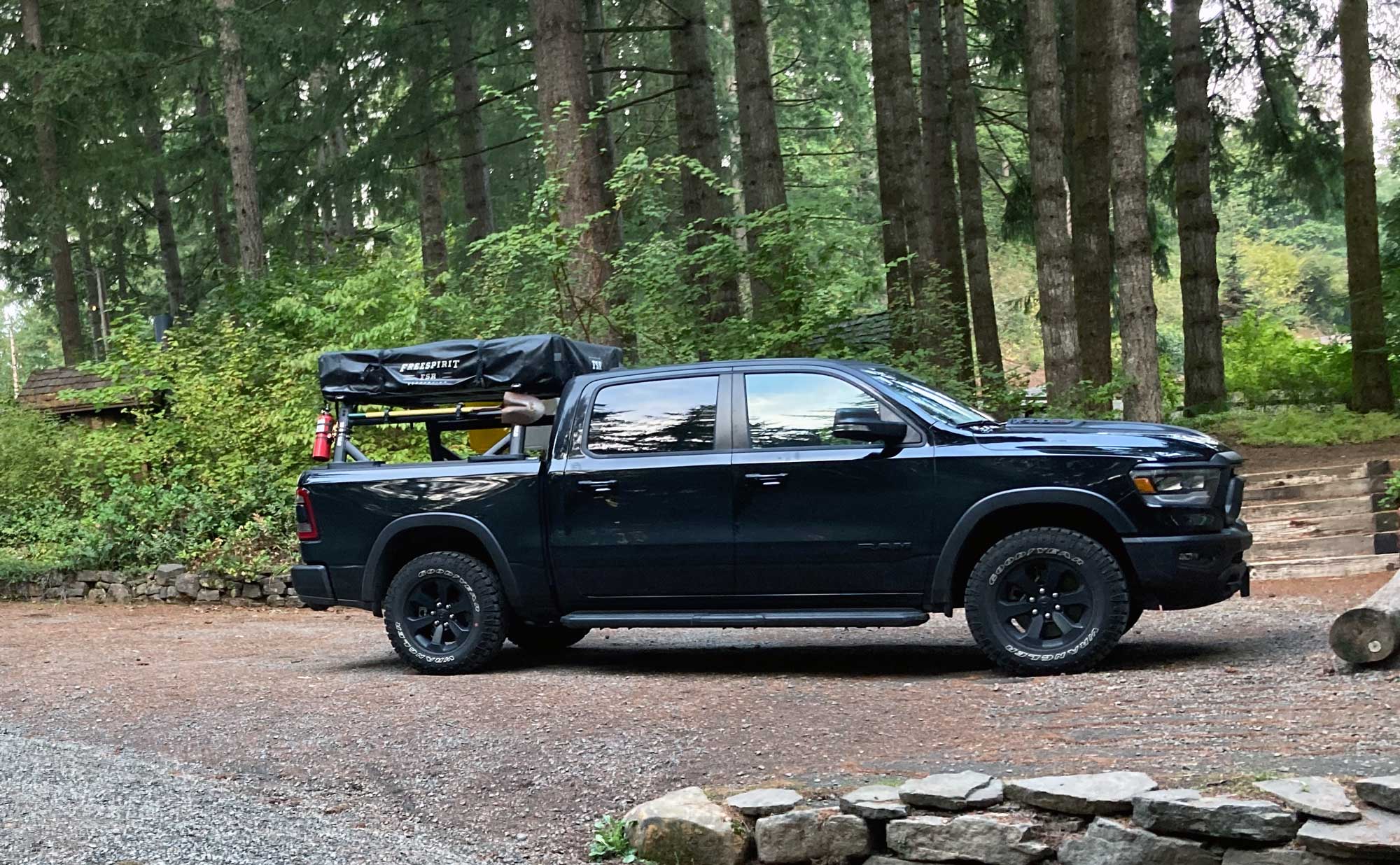 Black Dodge Ran four door truck with rack, roof top tent, shovel mount, and green trees in the background