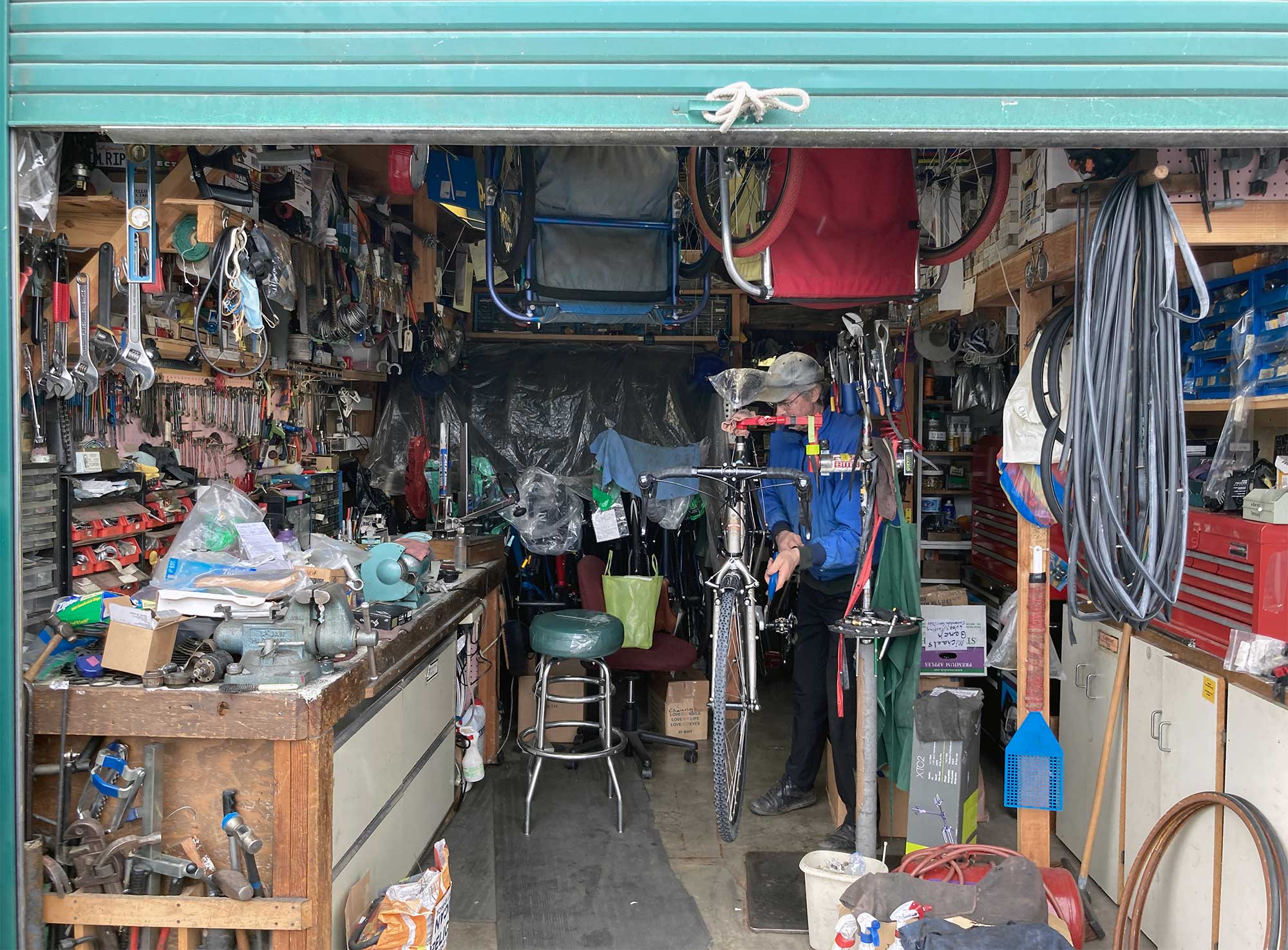 An open storage shed with a man working on a bike and bike tools, parts, gear, stools, and workbench around him
