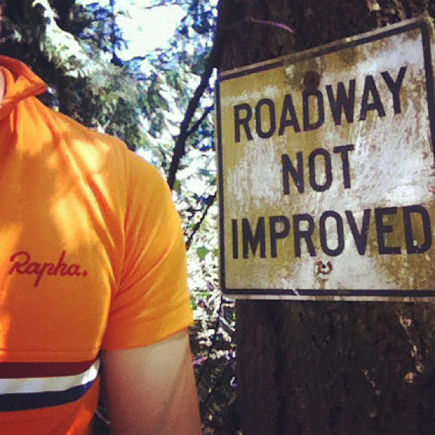 Left shoulder of a person with an orange shirt reading Rapha and a sign that reads Roadway not improved