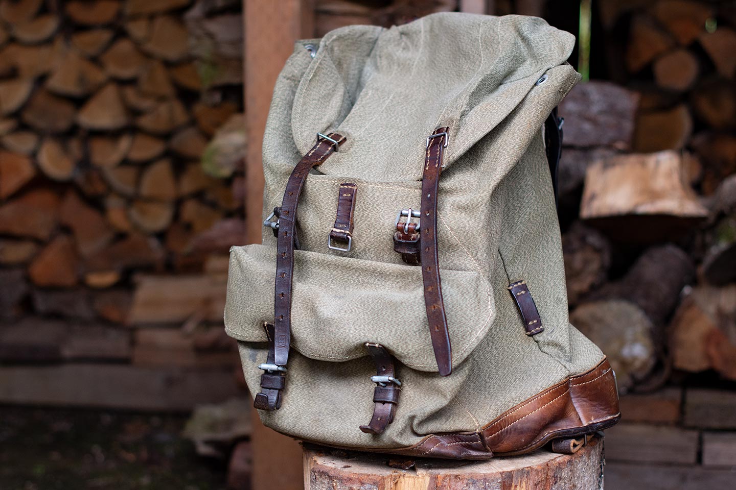 Canvas Swiss Army rucksack with leather fittings sitting on a stump in front of stacked wood