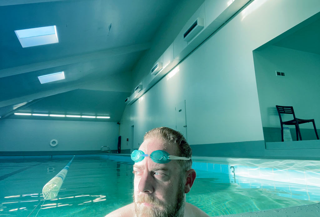 A person with a beard wearing light green goggles in a swimming pool with a high ceiling with skylights and two doors along one wall with a black chair in an alcove