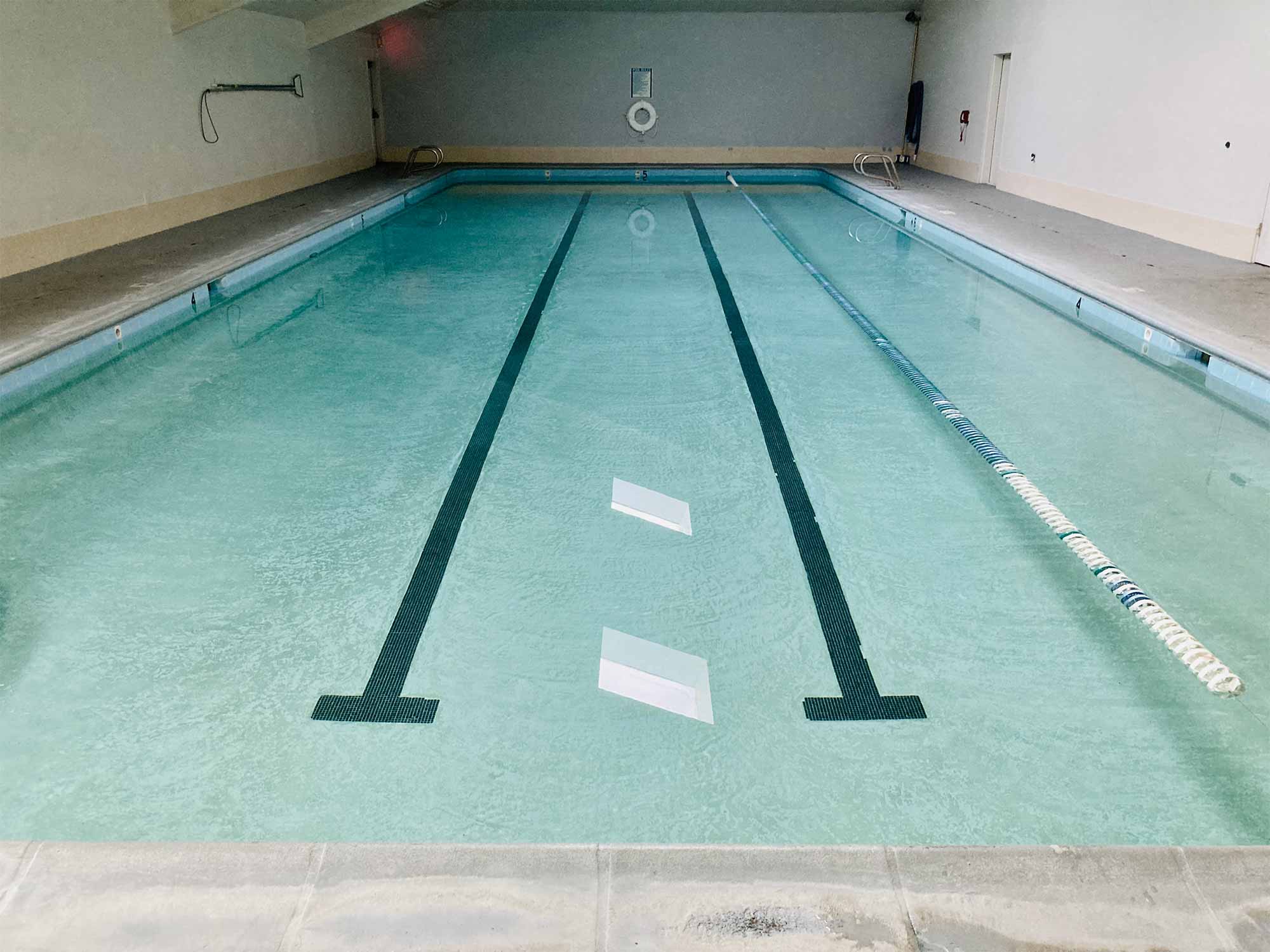 A worn swimming pool, with two distinct lanes with tile on the bottom and one lane line marking off the right side of the pool, grey and white walls and rescue and cleaning gear hung on the walls