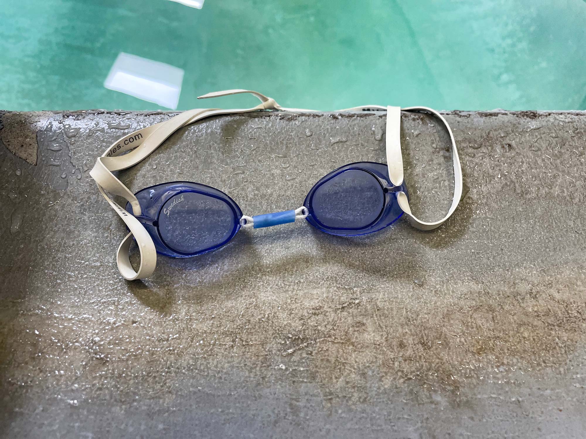 A set of violet goggles with a blue nose bridge and white band sit on a gray tile pool deck