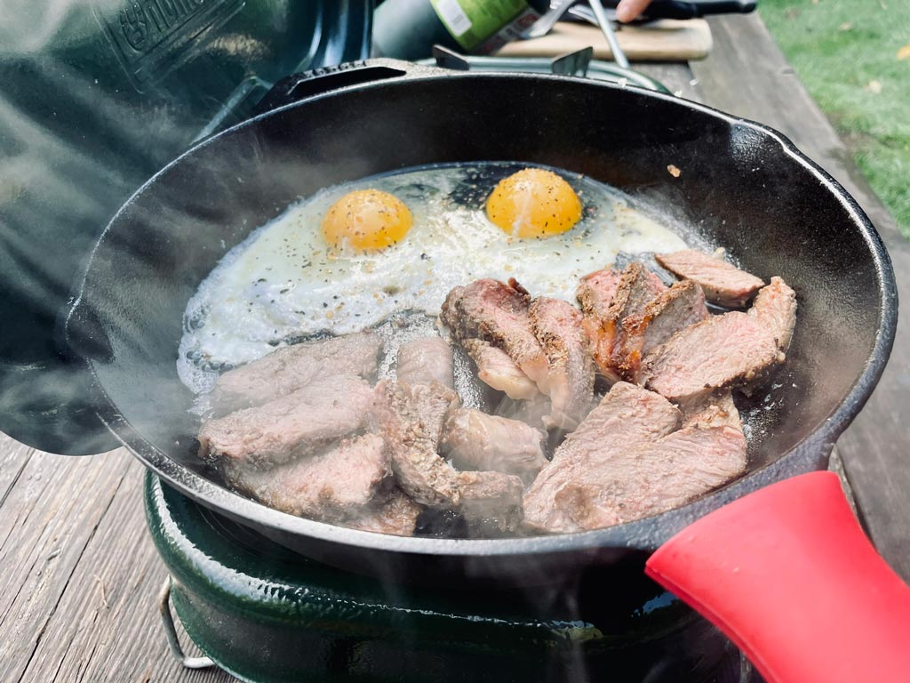 Slices of steak and two eggs steaming in a cast iron pan on a green propane stove