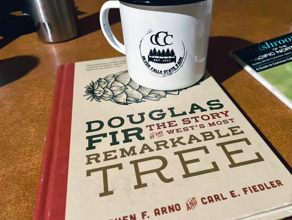 A white ceramic mug with CCC Silver Falls State Park established 1933 on the side sits on top of a book titled Douglas Fir the story of the west's most remarkable tree
