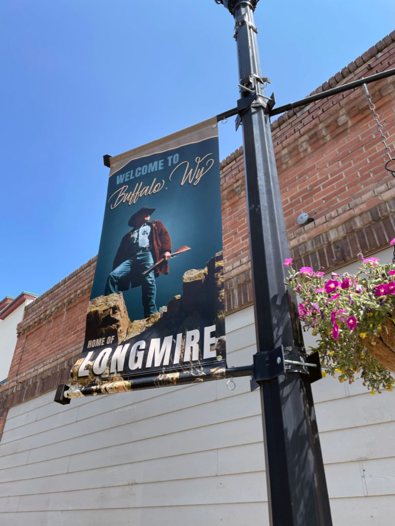 A banner reading Welcome to Buffalo, WY and Home of Longmire on a street light with a flower basket hanging