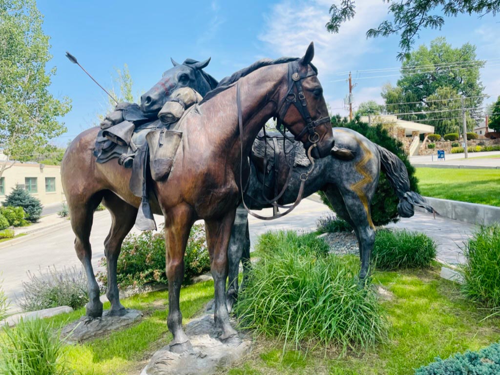 A bronze sculpture of two horses, one resting its head on th other, and an arrow protruding from a pack on the back of one horse; the sculpture is surrounded by plants, roads, and buildings in the background