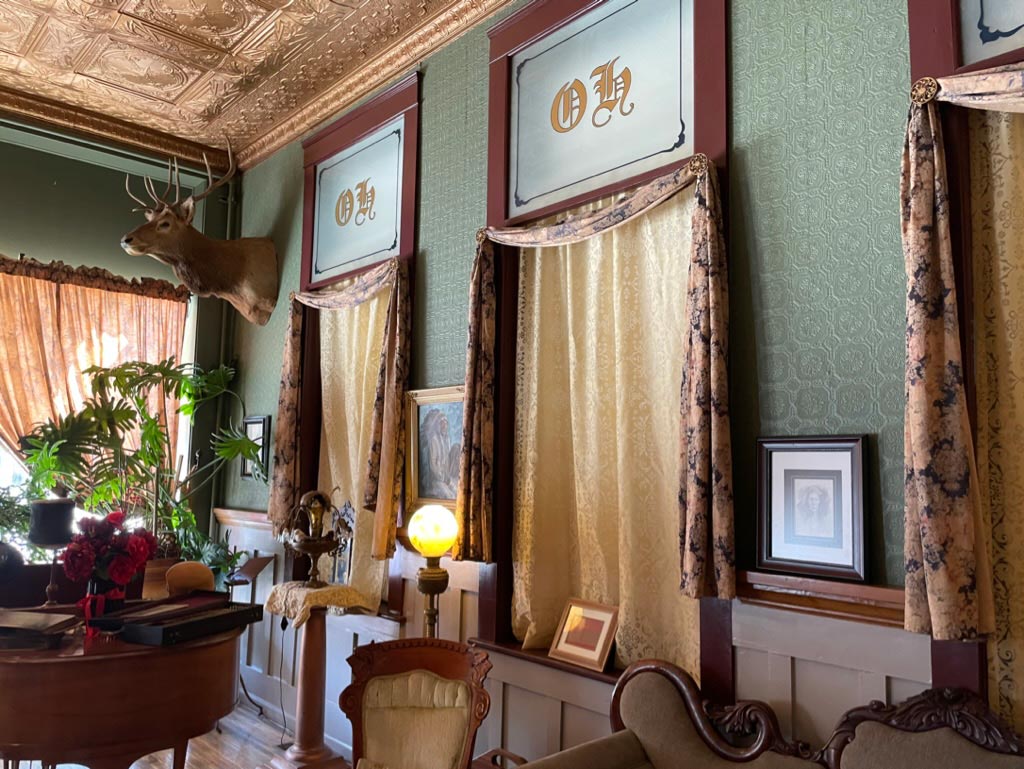A taxidermied elk head with windows and drapes, wood carved furniture, elegant sea-foam wallpaper, and framed pictures on the walls