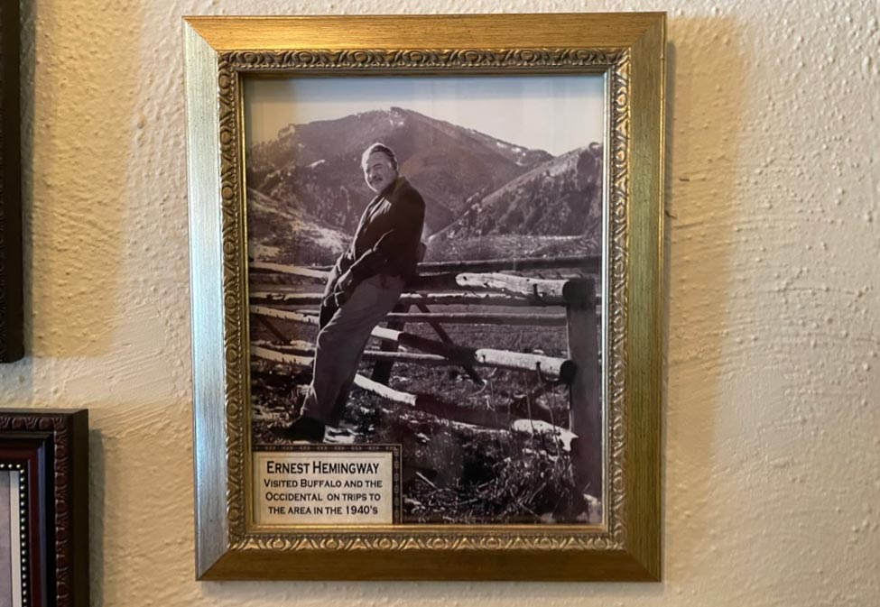 A framed picture of Ernest Hemingway leaning on a wooden fence with mountains in the background and a note that reads Ernest Hemingway listed Buffalo and the Occidental on trips to the area in the 1940s