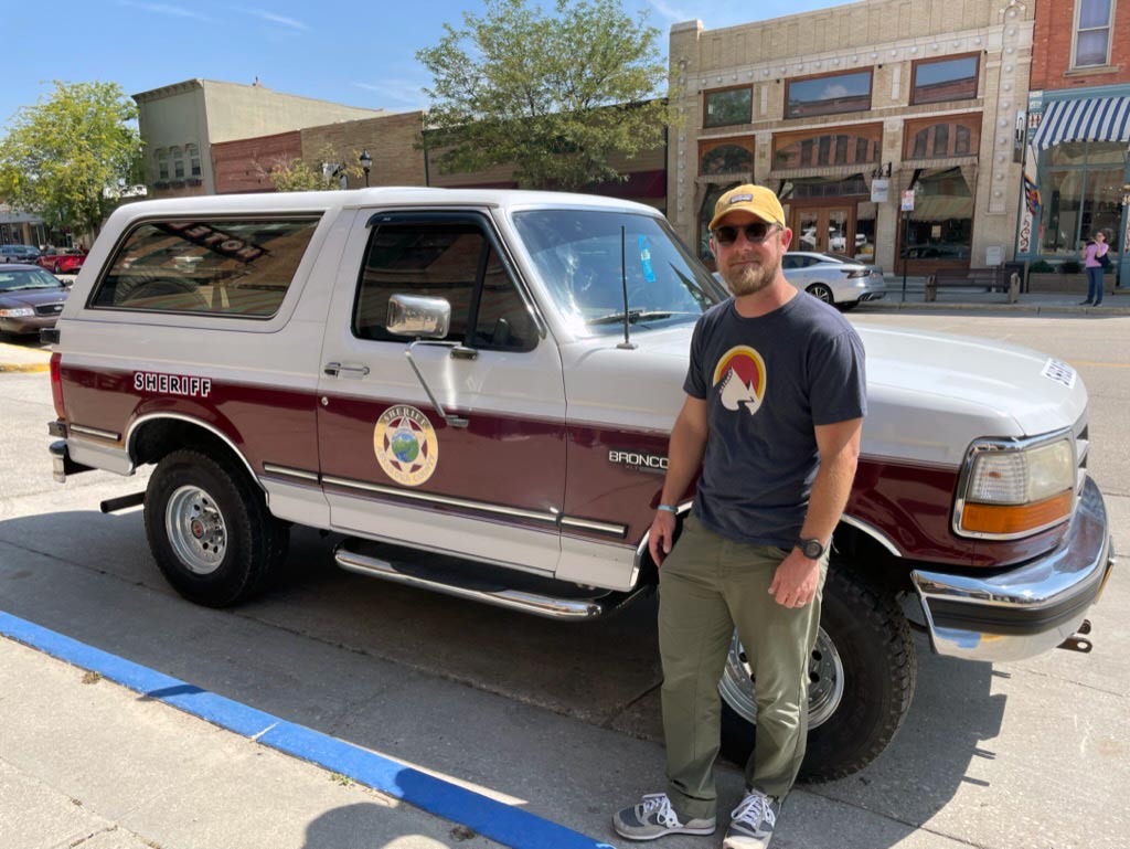 Wearing a yellow hat, blue shirt, and green pants with sunglasses, I stand next to a white Ford Bronco with a maroon stripe not the side and a gold star that reads Sheriff Absaroka County