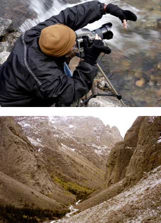 A person takes a photo near a river and blocks the sun with their left hand, the person is wearing a black jacket and tan beanie hat; below that, a snow lined mountain landscape with a river running through