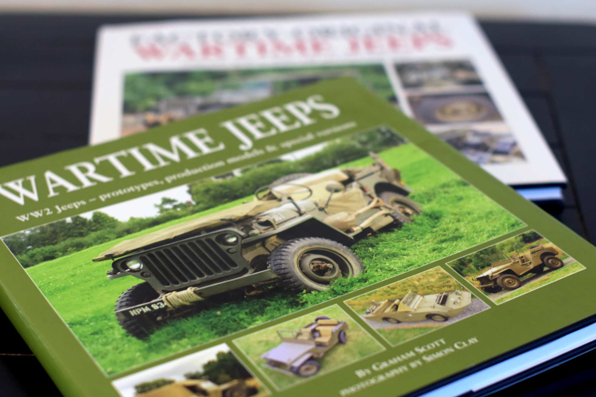 Two books, one out of focus vaguely with the title Wartime Jeeps, and the in-focus book in green also with the title Wartime Jeeps and WW2 jeeps also visible with a vehicle, the jeep, below in olive drab paint