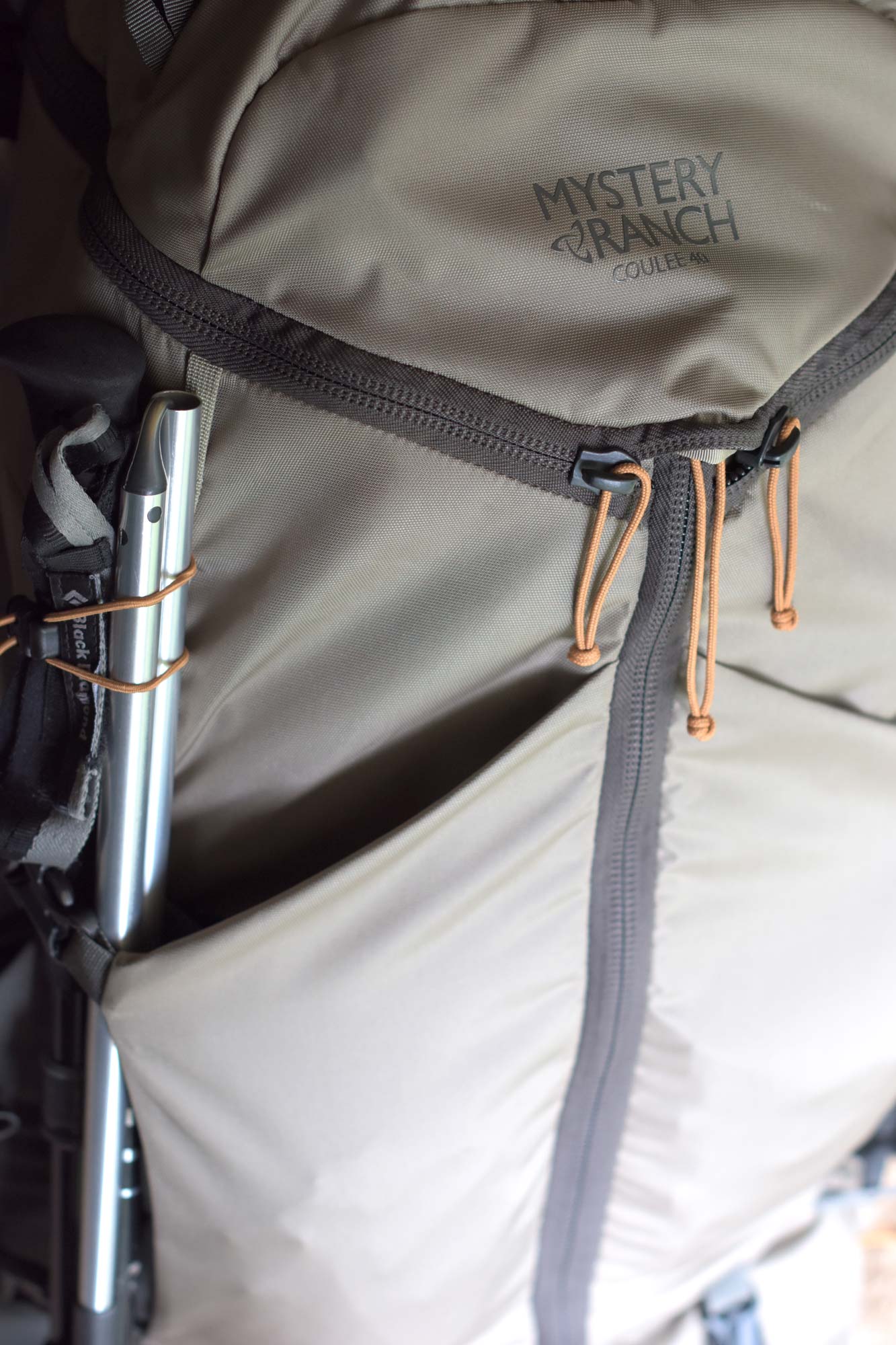 A tan backpack with Mystery Ranch Coulee 40 in black with black zippers, orange zipper pulls, and a trekking pole lashed to the left side