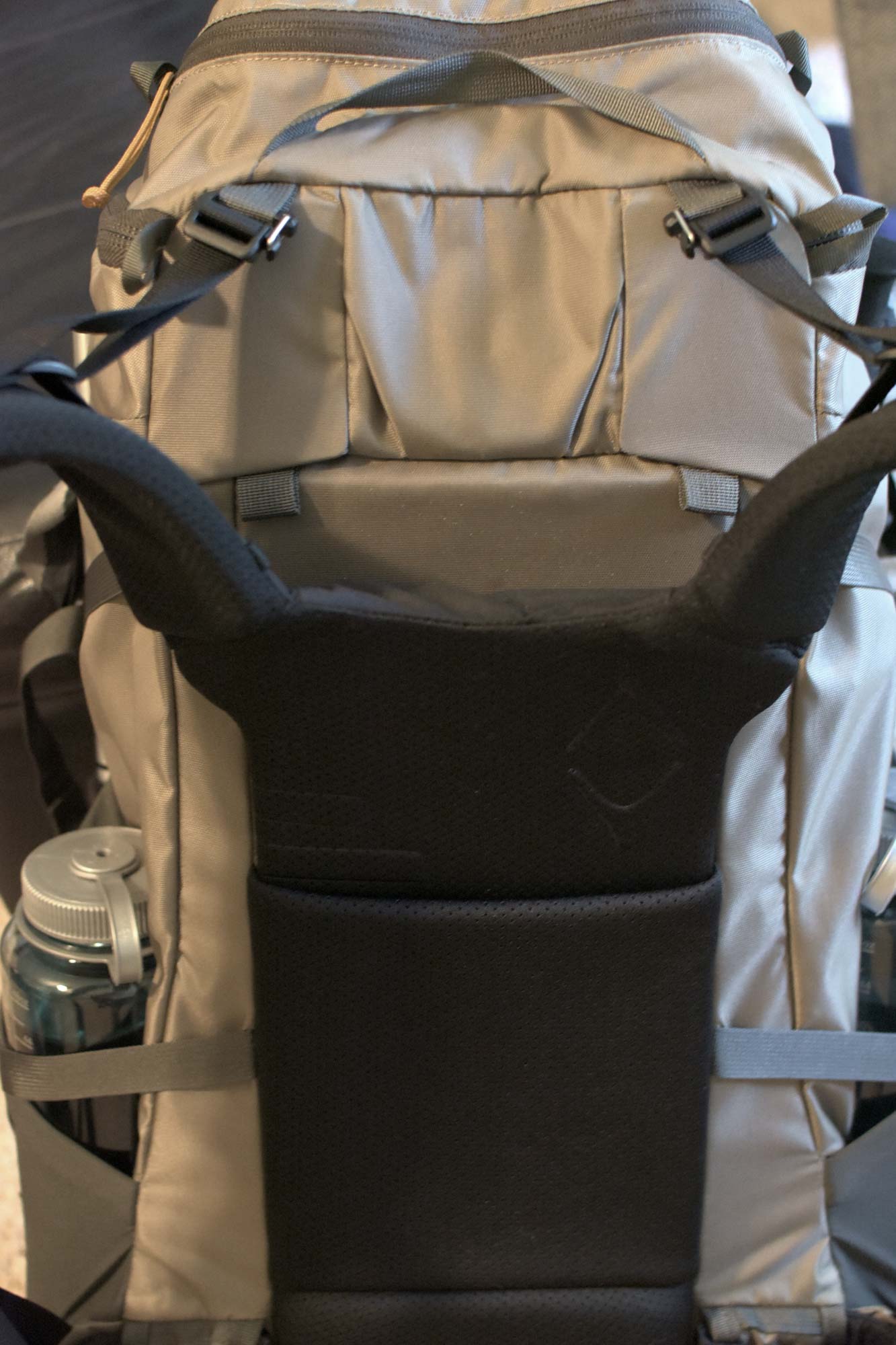 A tan backpack from the back showing black shoulder straps and a black back padding with a water bottle in the left pocket