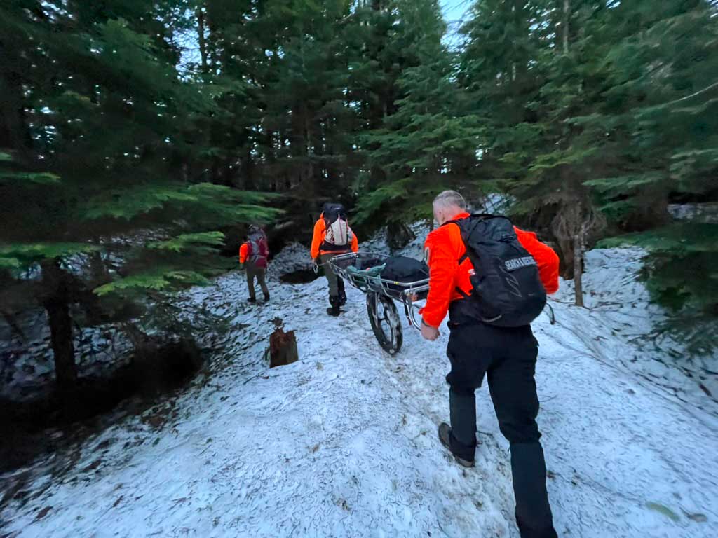 Search and rescue team members dressed in orange guide a wheeled litter basket along a snowy trail with trees lining the trail on each side