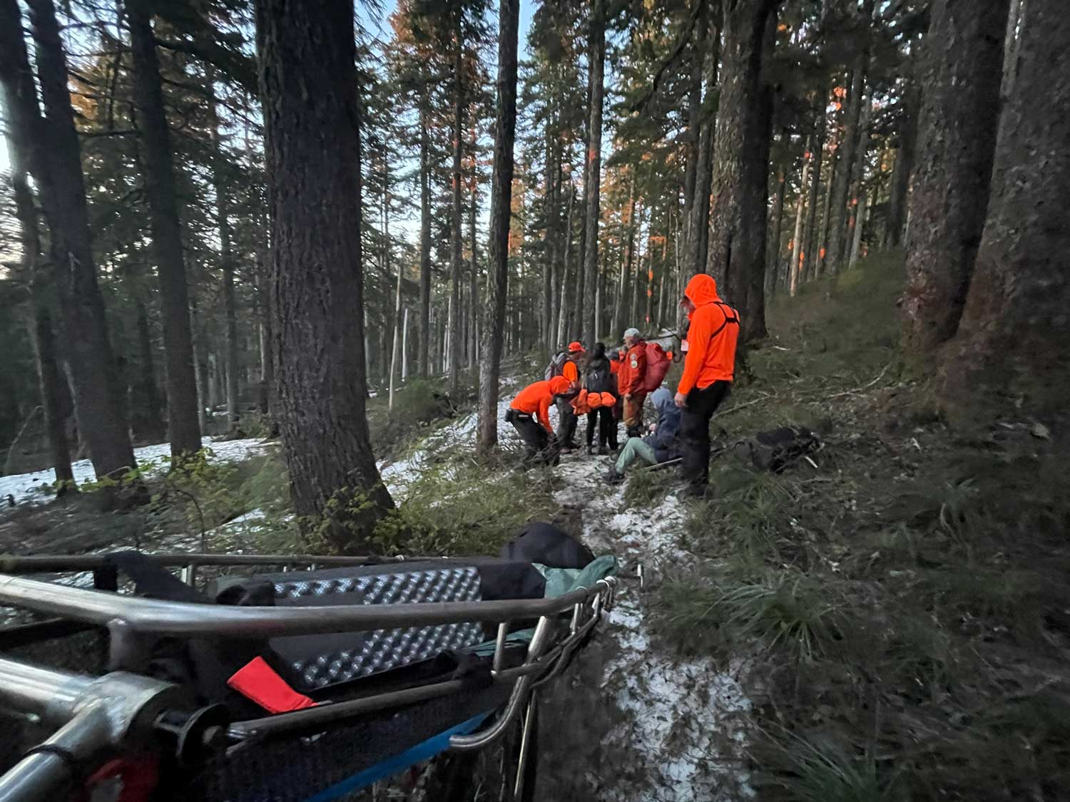 Several search and rescue team members wearing orange gather around someone sitting on the ground along a trail, with a litter basket in the foreground