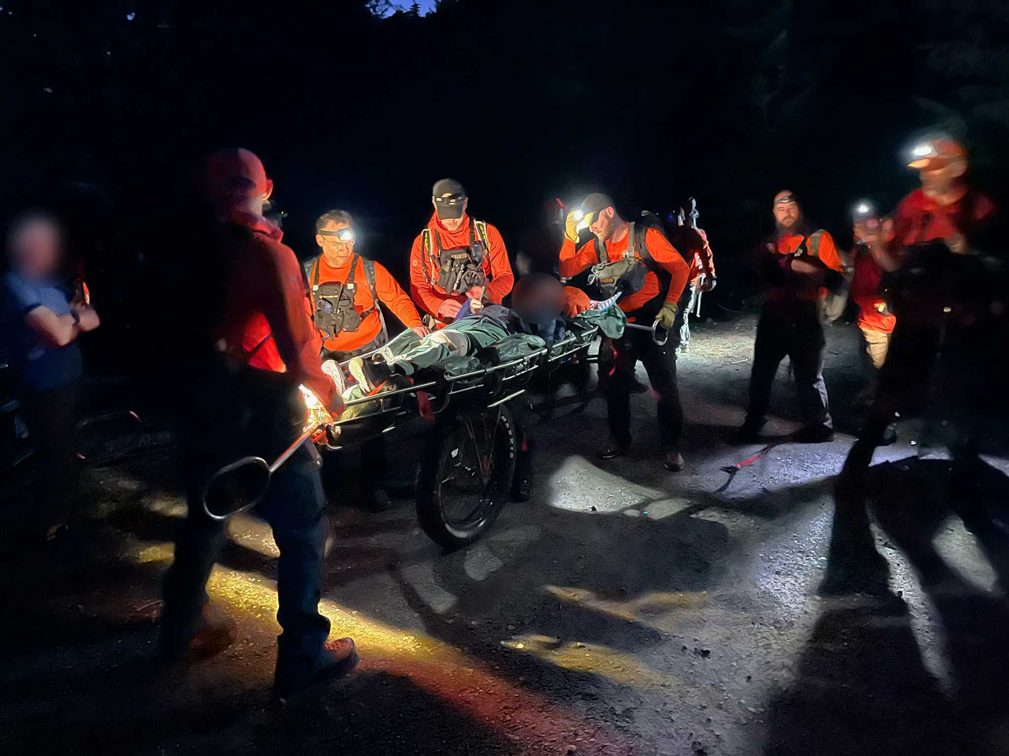 Several search and rescue team members wearing orange shirts surround and secure a wheeled litter bearing a person; it is nighttime and several of the team members wear headlights in the dark, casting light and shadow around the scene