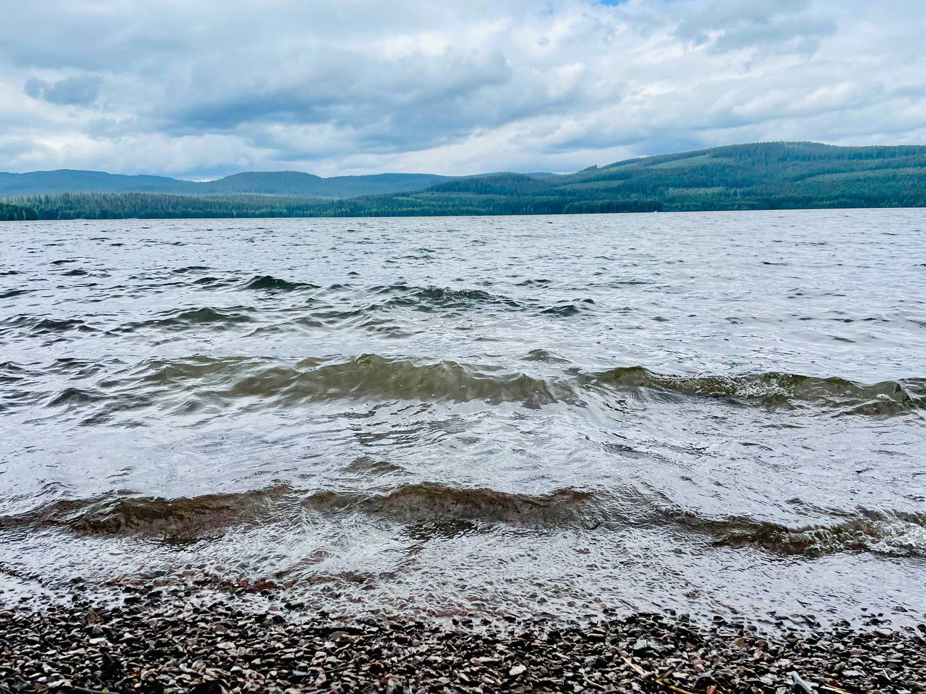 A cloudy sky with green hills in the distance lining a lake surface with choppy waves and a gravel shore in the foreground