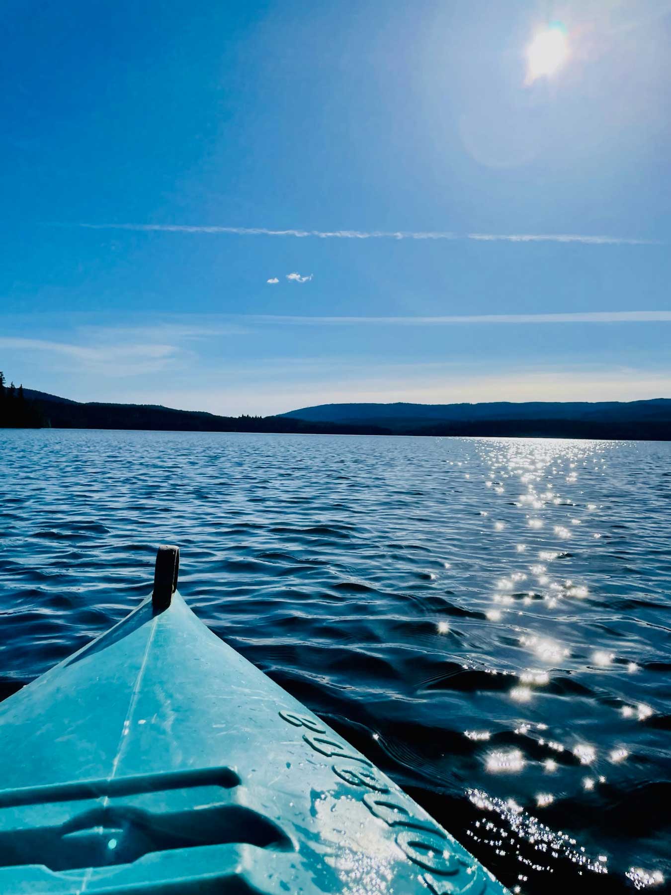 The front of a turquoise kayak out on a lake with the sun shimmering off the lake surface and hills in the distance