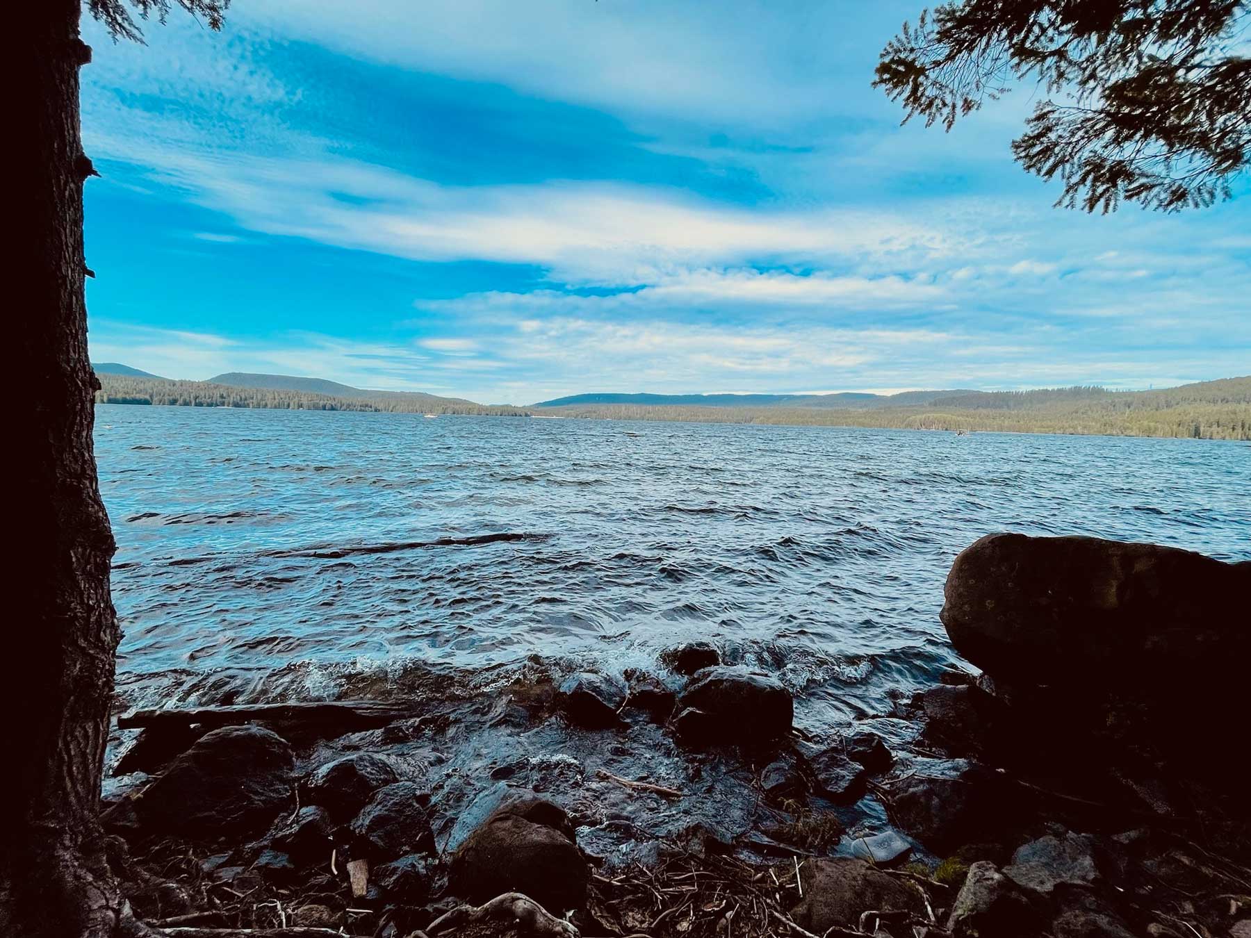 A lake with moderate waves washing ashore framed by trees with the far tree-lined, lakeshore hills in the distance