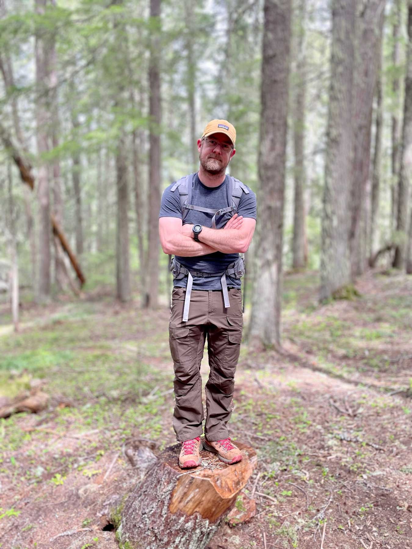 A man crossing his arms wearing a backpack, yellow-orange hat, gray shirt, brown pants, and hiking boots stands on a stump among trees in a forest