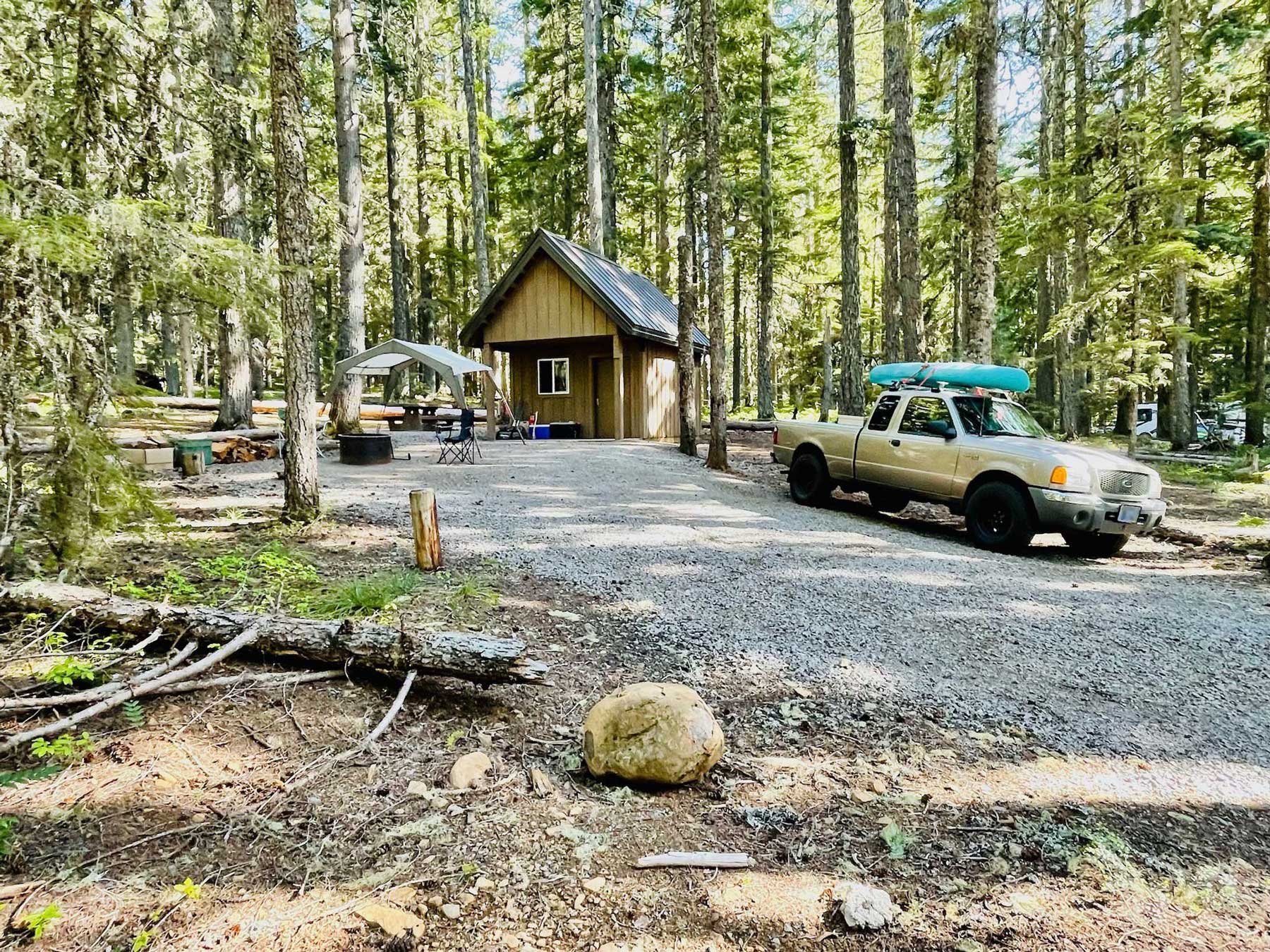 A small cabin among trees with a gravel driveway and a tan pickup truck with a turquoise kayak on top