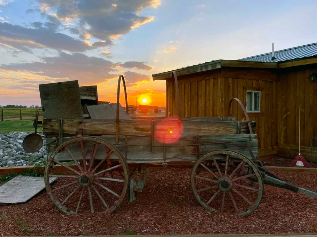 An old pioneer wagon sits among red bark chipped ground in front of a rustic building, with an orange sun setting the background