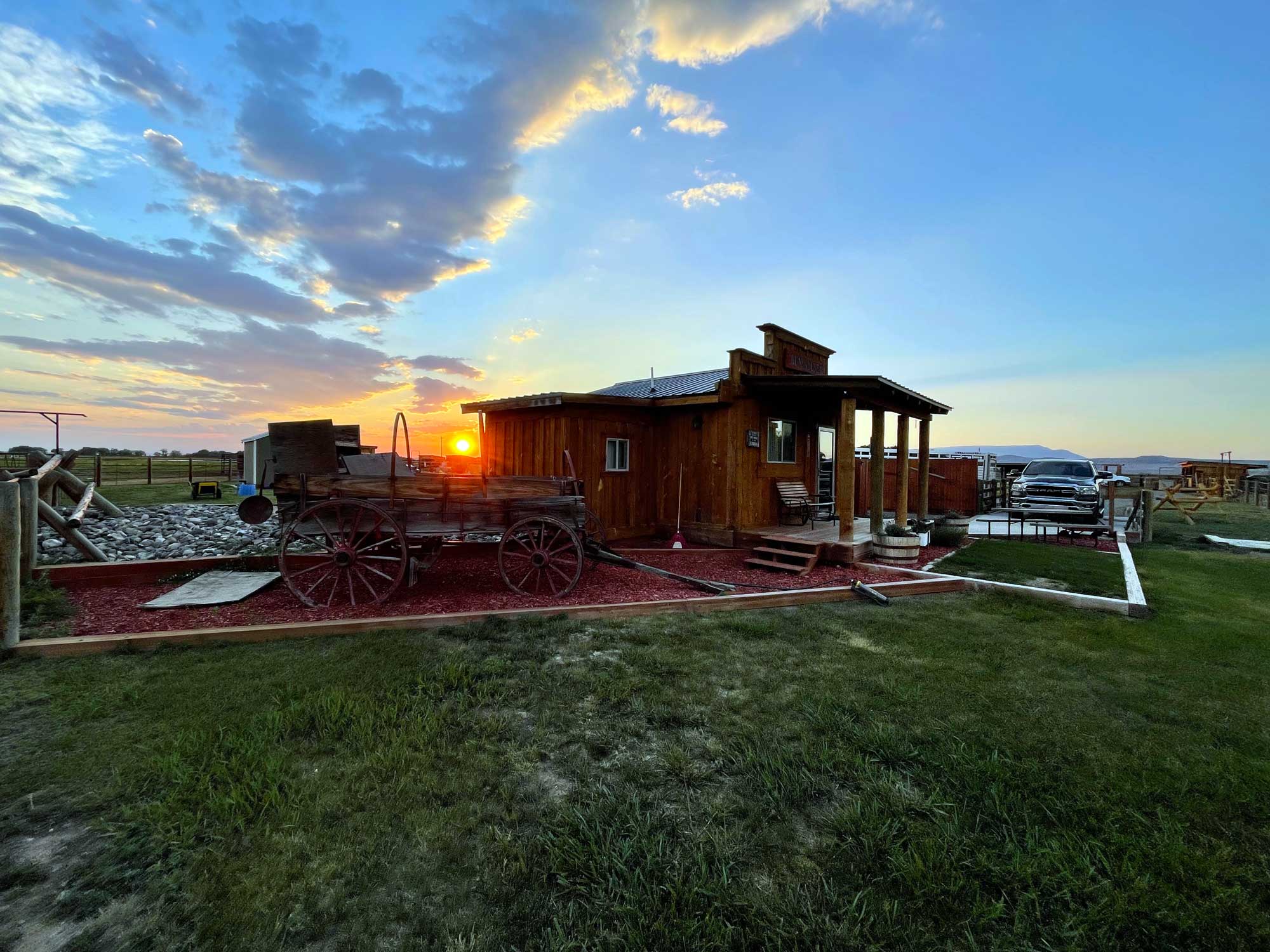 An orange sun sets behind an old horse-drawn wagon and a rustic bunkhouse, with a silver Ram truck on the right side and trimmed grass in the foreground