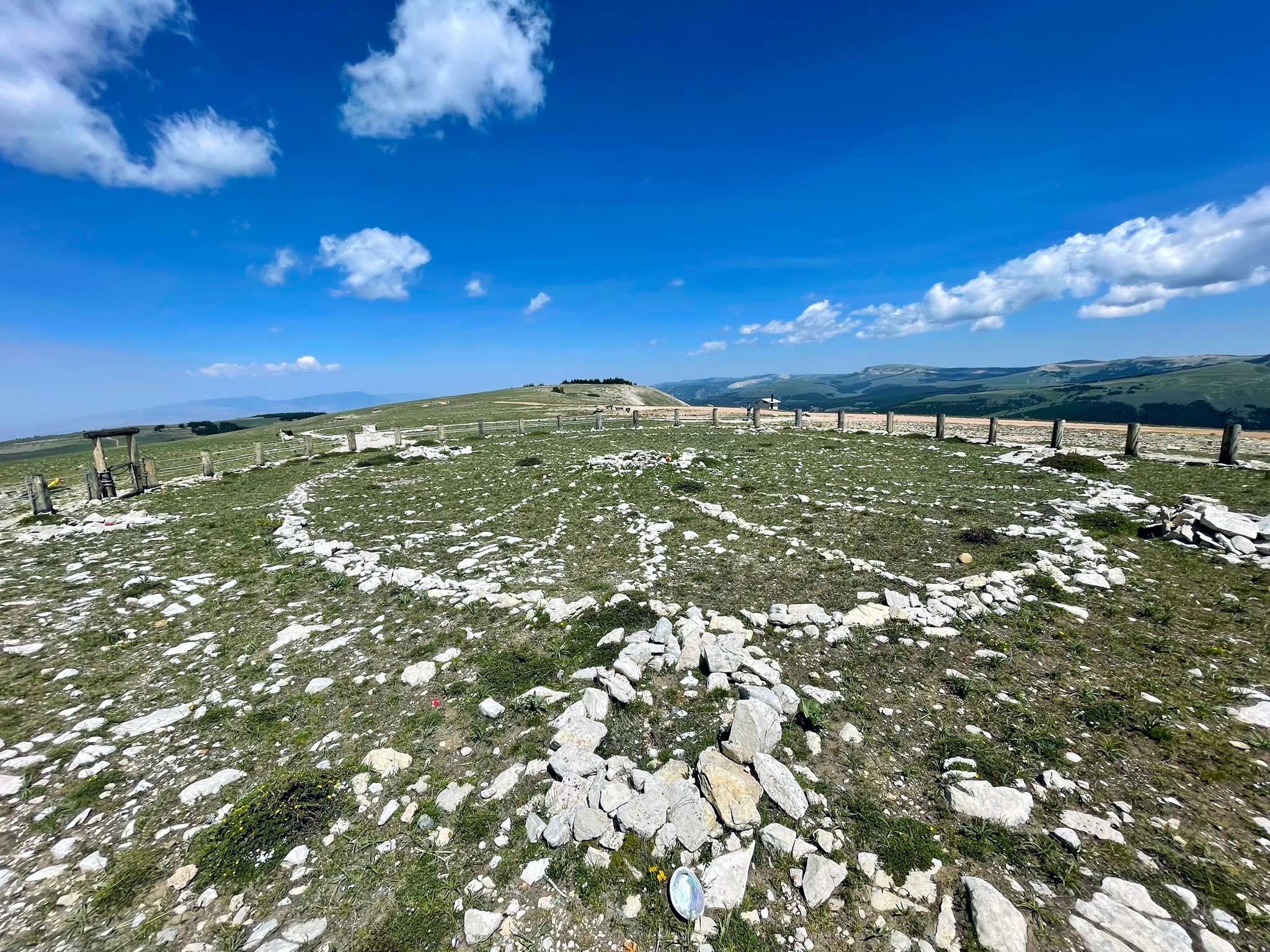 The medicine wheel, a circular spiral of light colored rock with fence posts surrounding it, with green mountain slopes in the background, and a blue sky with a few white clouds overhead