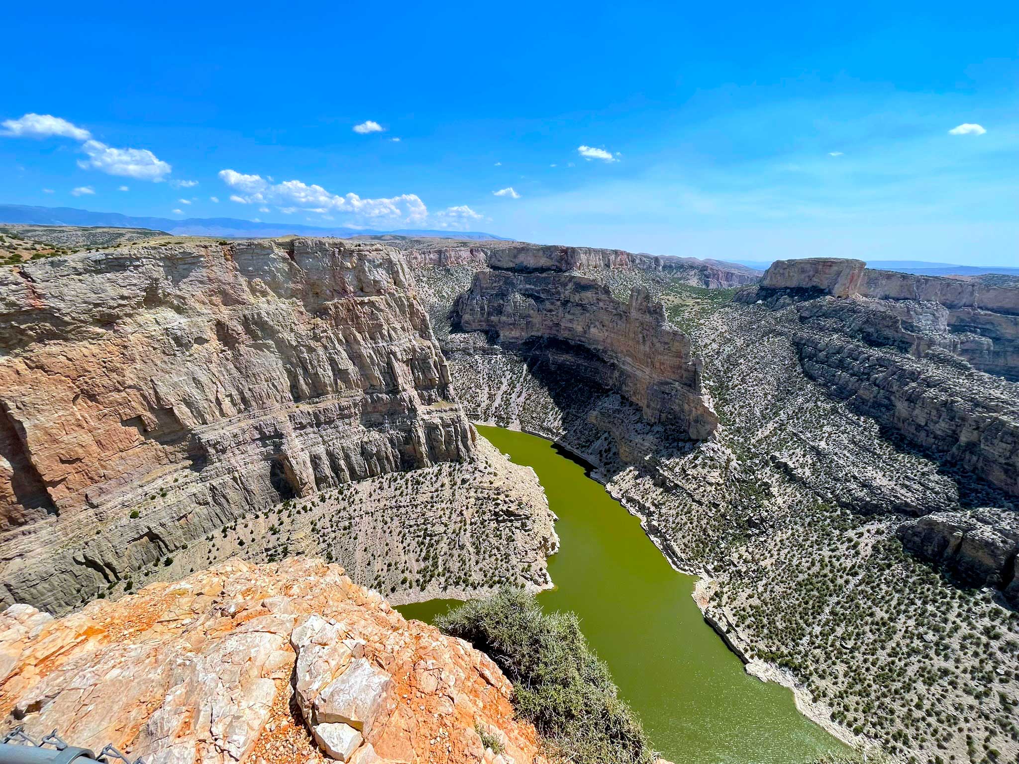 A deep rocky canyon with multiple colors of rock, dotted with desert shrubs, with a winding green river at the bottom of the canyon