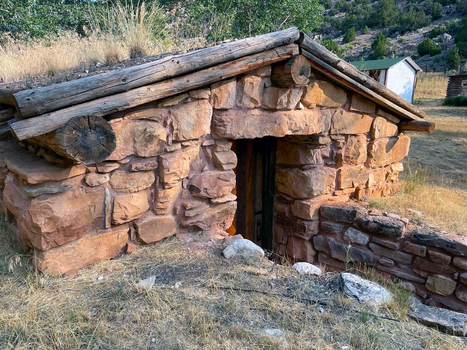 A reddish-yellow-colored stone building with a door that extends below the ground level, with log roof framing