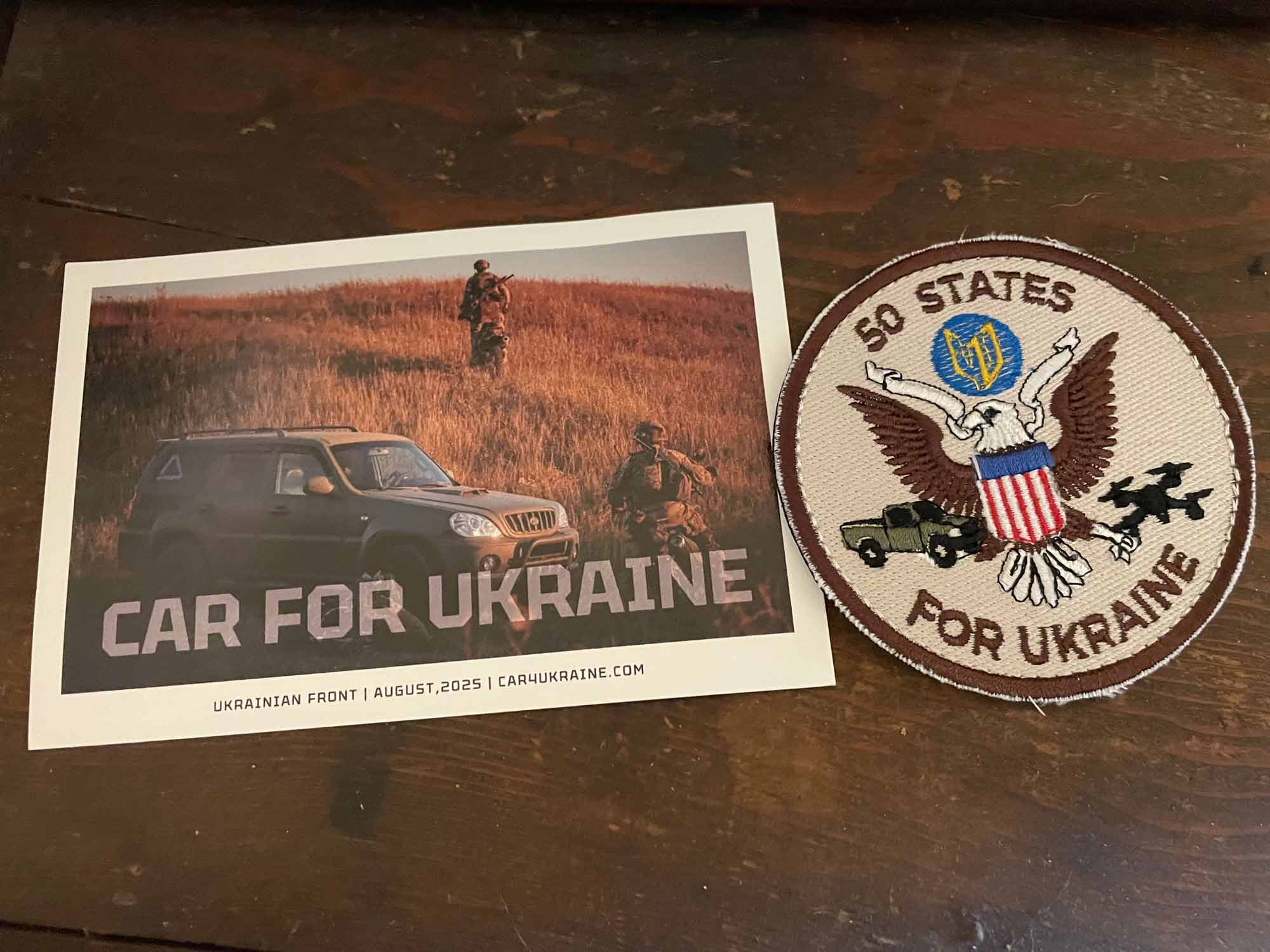 A postcard shows two military troops near a small armored SUV; the card reads Car for Ukraine, Ukrainian Front, August 2025, car4ukraine.com; next to the postcard is a patch with an eagle, USA and Ukraine symbols, a pickup truck and drone, and the text 50 states for Ukraine