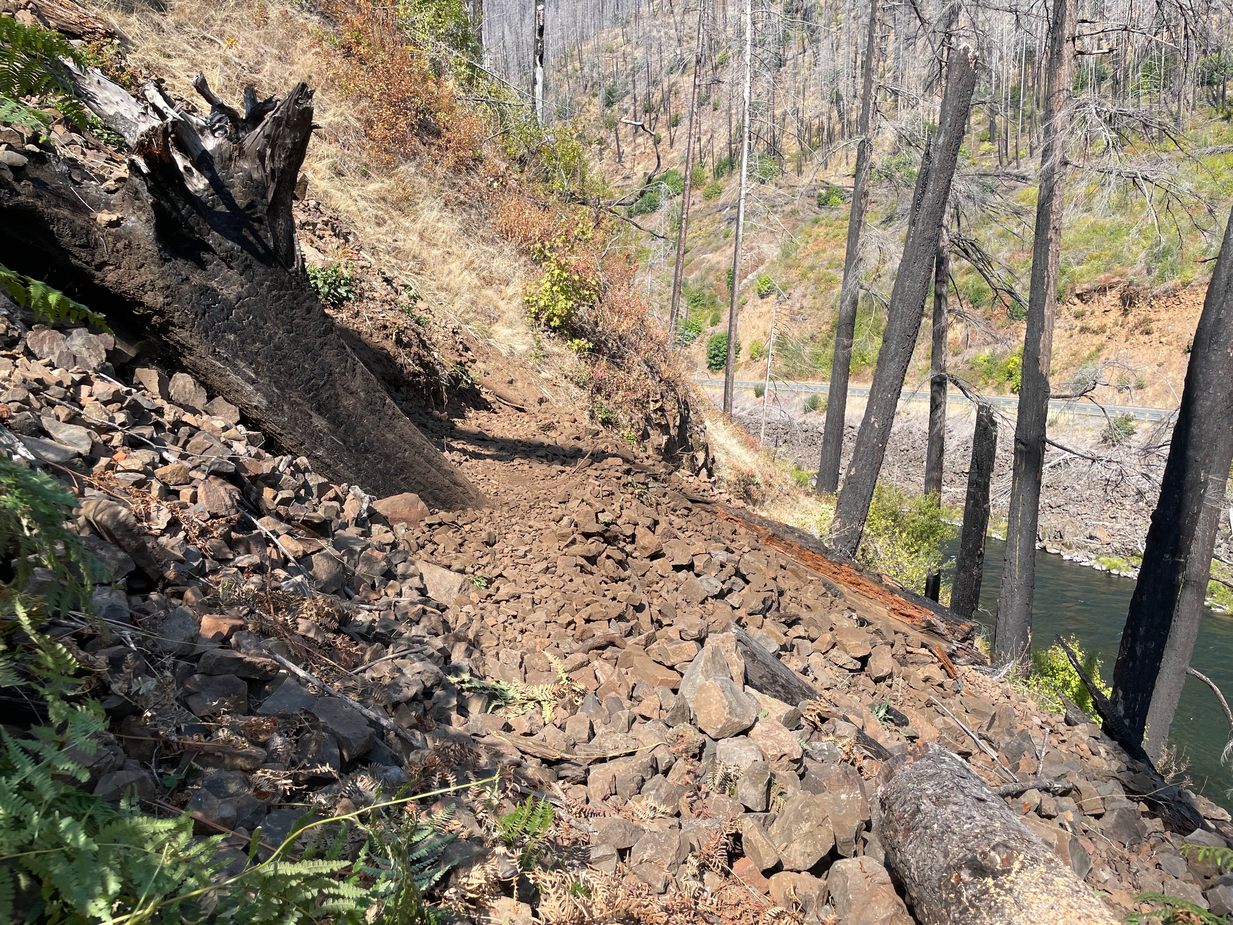 A faint trail across a rockslide with burned trees on the hillside