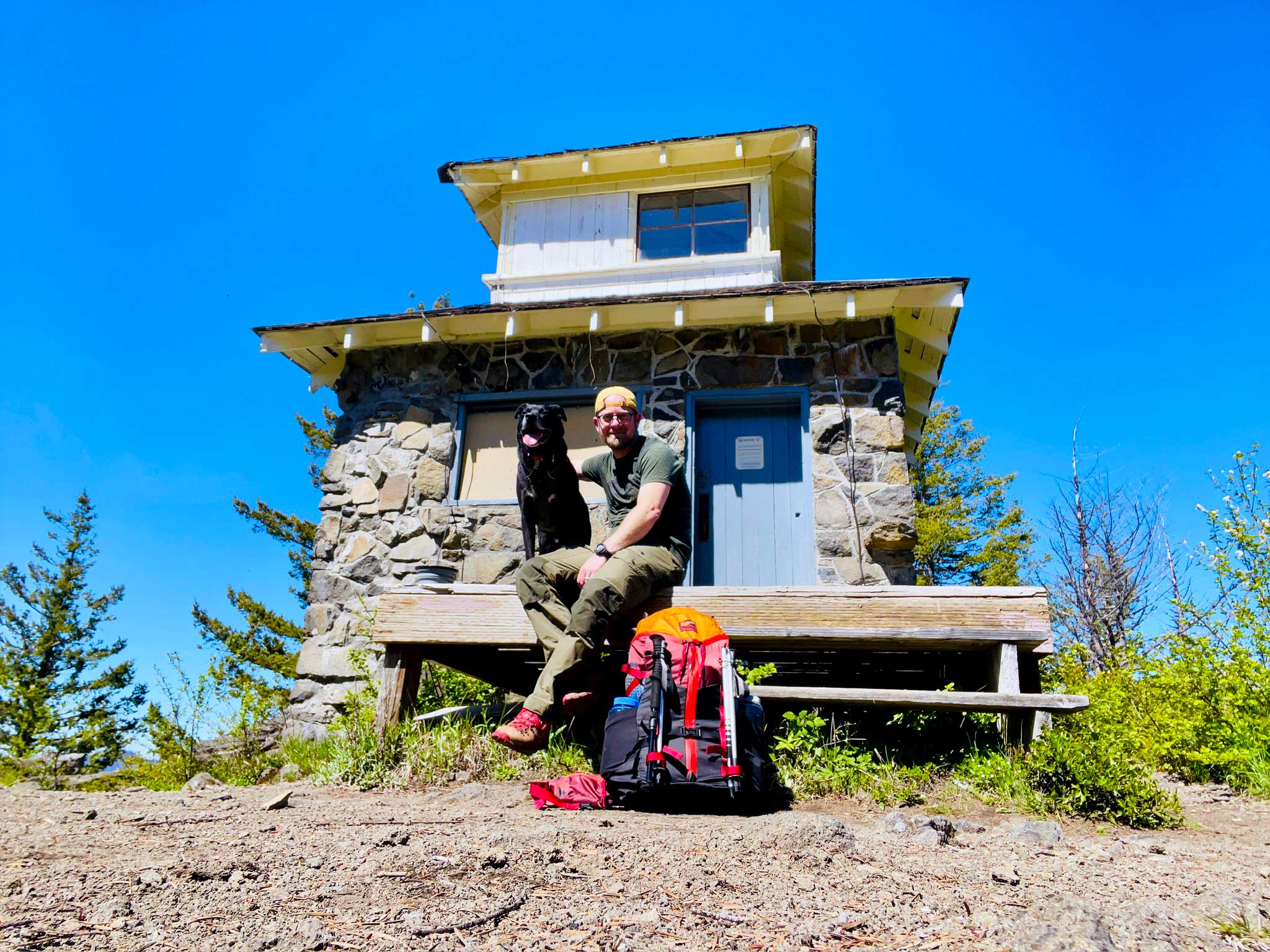 A man wearing a yellow hat backwards, green shirt and pants, and hiking boots sitting with a panting black dog on the porch of a small stone building with a blue door with blue sky and trees in the background, there's an orange and red backpack in the foreground