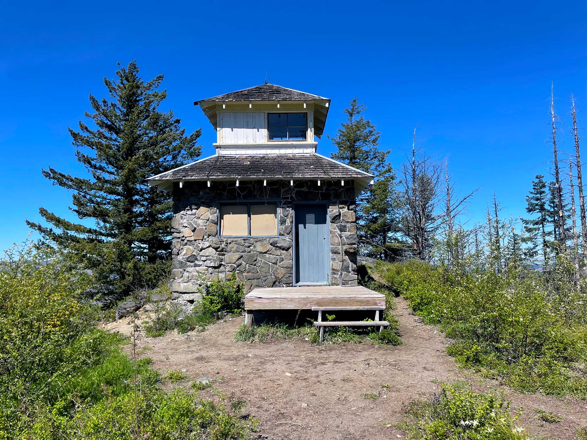 A stone fire lookout building with a blue door and porch with green trees and some burned trees around