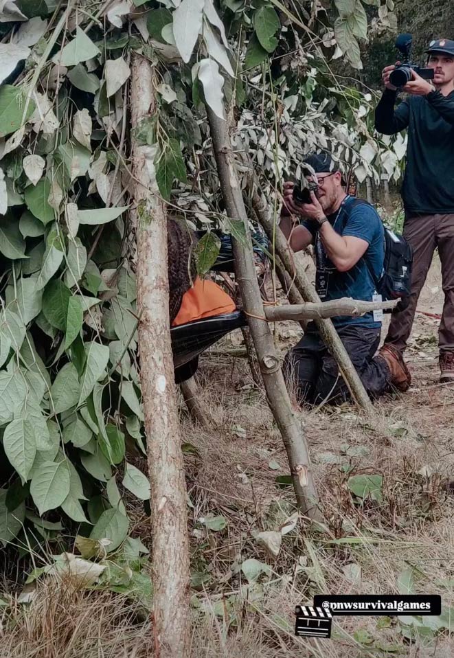 Two photographers pointing cameras toward a person in an orange suit lying in a shelter made of branches and leaves