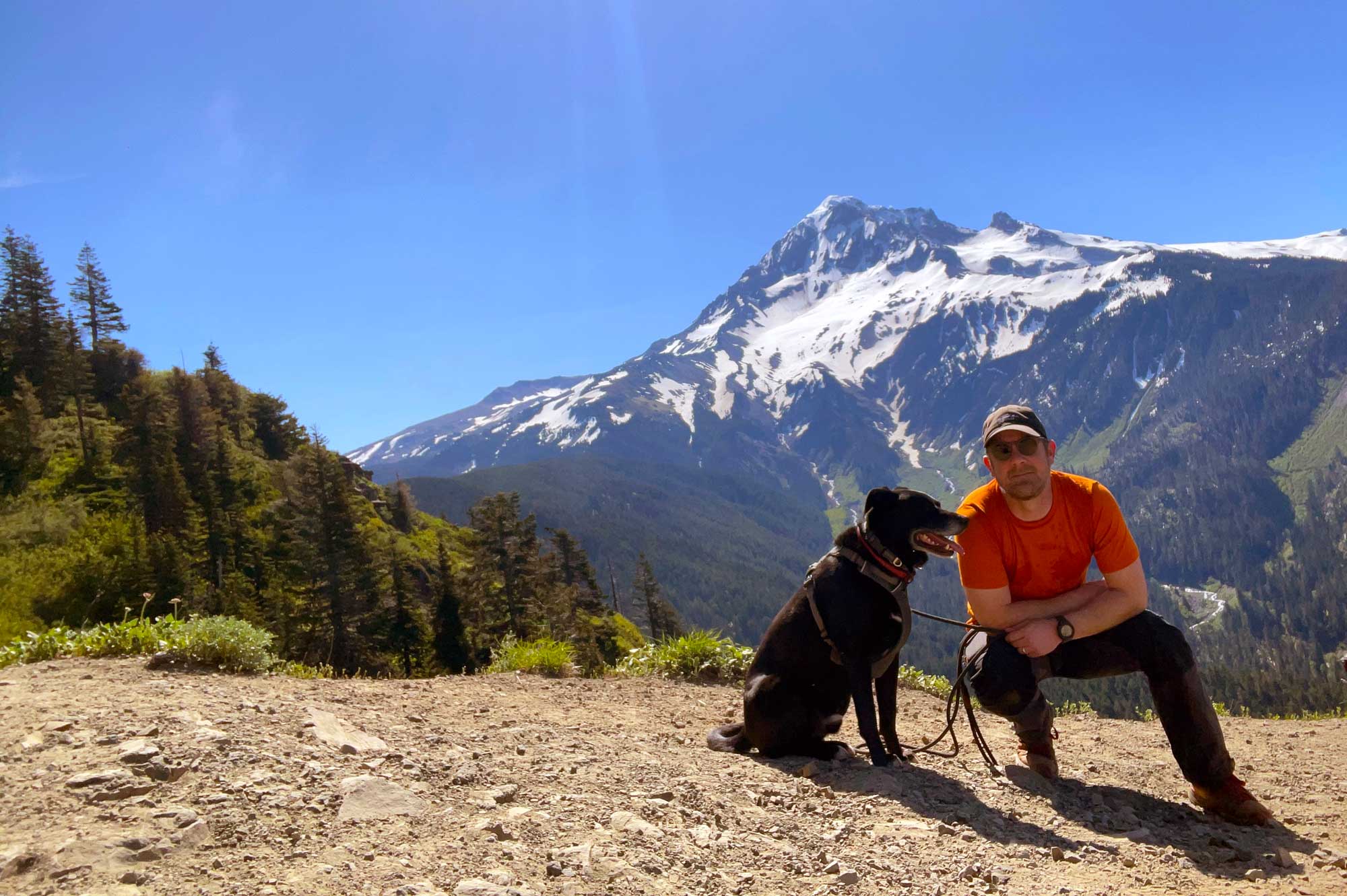 A man wearing an orange shirt and black cap with a black dog beside him on a rocky trail with a snow capped mountain in the background