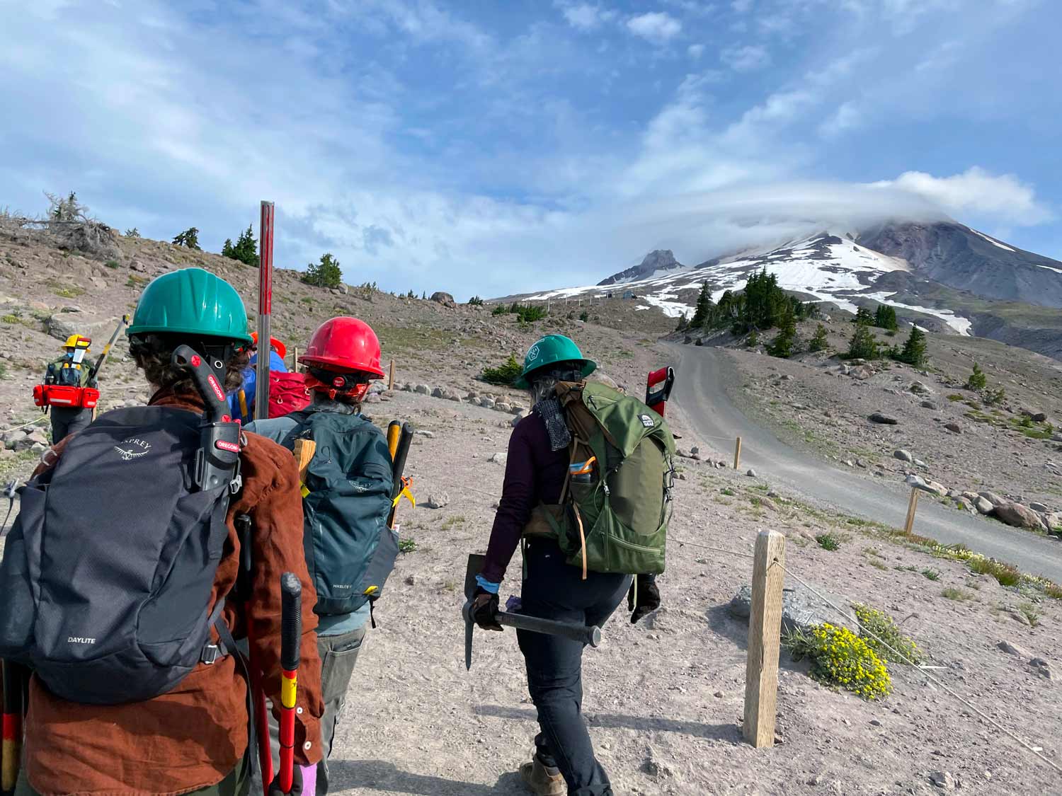 People in green, red, and yellow hard hats walk with backpacks on and holding tools along a gravel path at the base of a snow capped mountain with clouds covering the summit