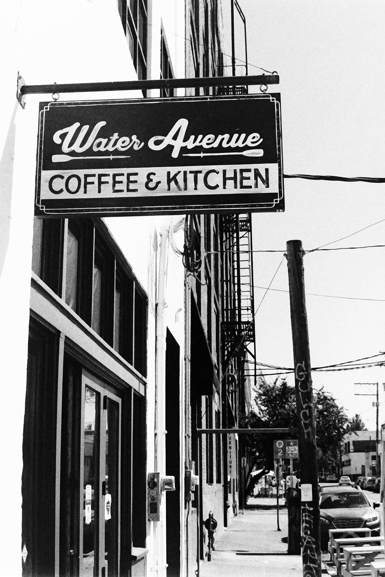 The sign for water avenue coffee and kitchen with the sidewalk below and a power pole with graffiti nearby