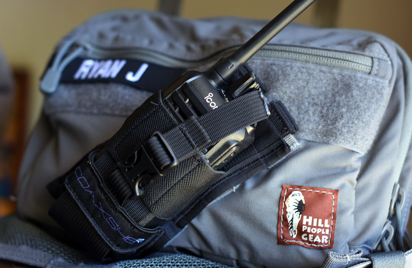 Gray Hill People Gear chest bag with radio harness with an iCom radio attached, and a name badge that reads Ryan J with a Hill People Gear rust colored badge at lower end of the bag
