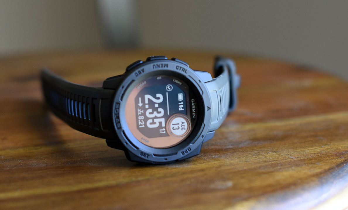 Gray and black Garmin Instinct watch sitting sideways on a wooden surface reading August 13 2:35 and 19 days of battery life with a horizontal arrow and sunset icon with 8:21 listed, and the words on the dial reading ctrl, menu, abc, solar, gps, and set
