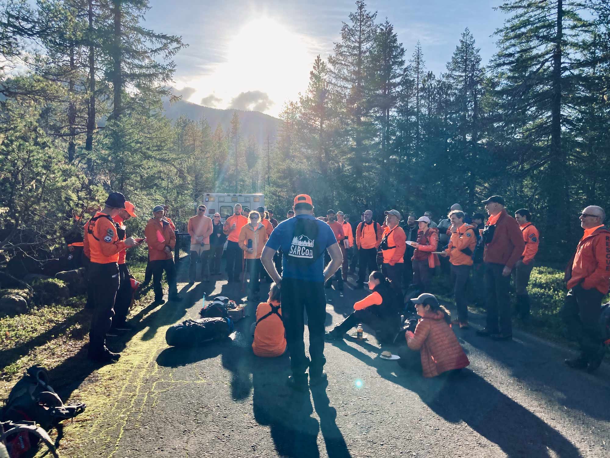 Several search and rescue members wearing orange shirts, some wearing blue, gathered together on a campground road in the forest